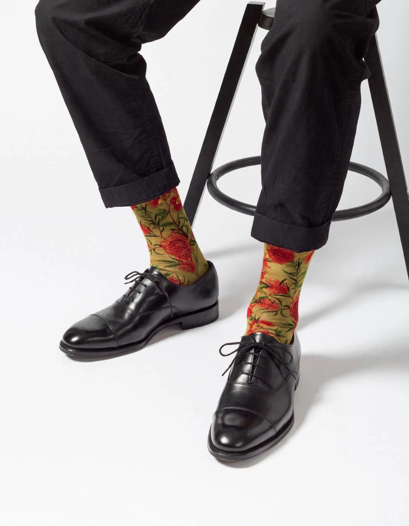 A person seated on a black stool wearing black trousers, black leather oxford shoes, and bright gumnuts flower patterned socks with red flowers and green leaves on a yellow background.