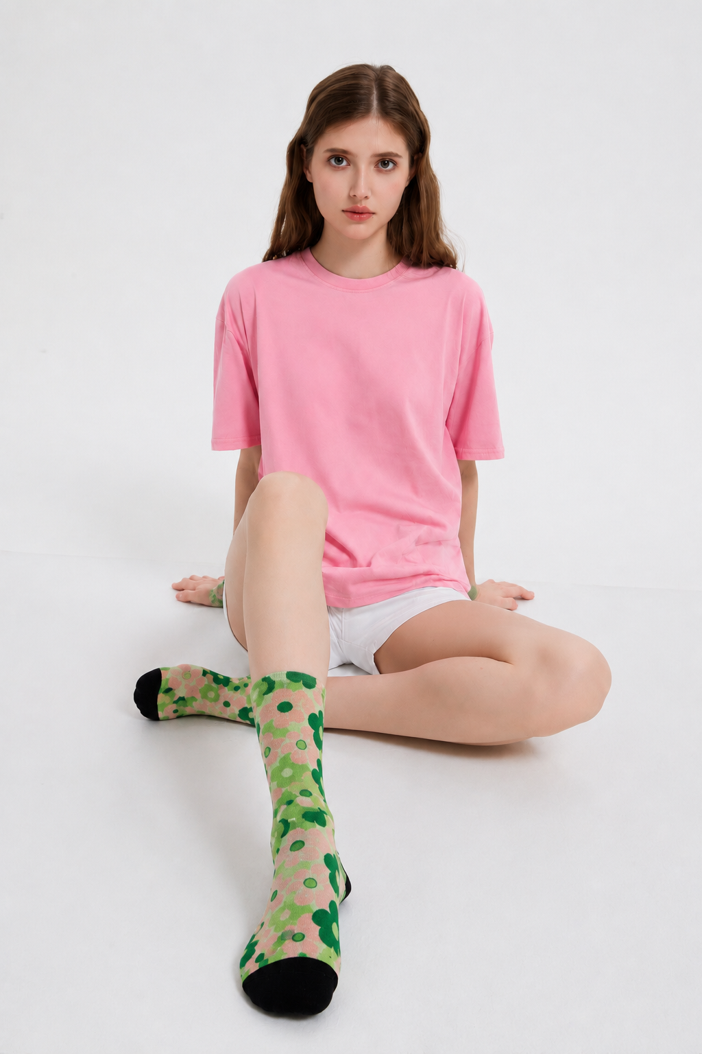 A model sitting on a white studio floor wearing a pink t-shirt, white shorts, and a pair of green and pink patterned Nordic flower socks with black toes and heels.