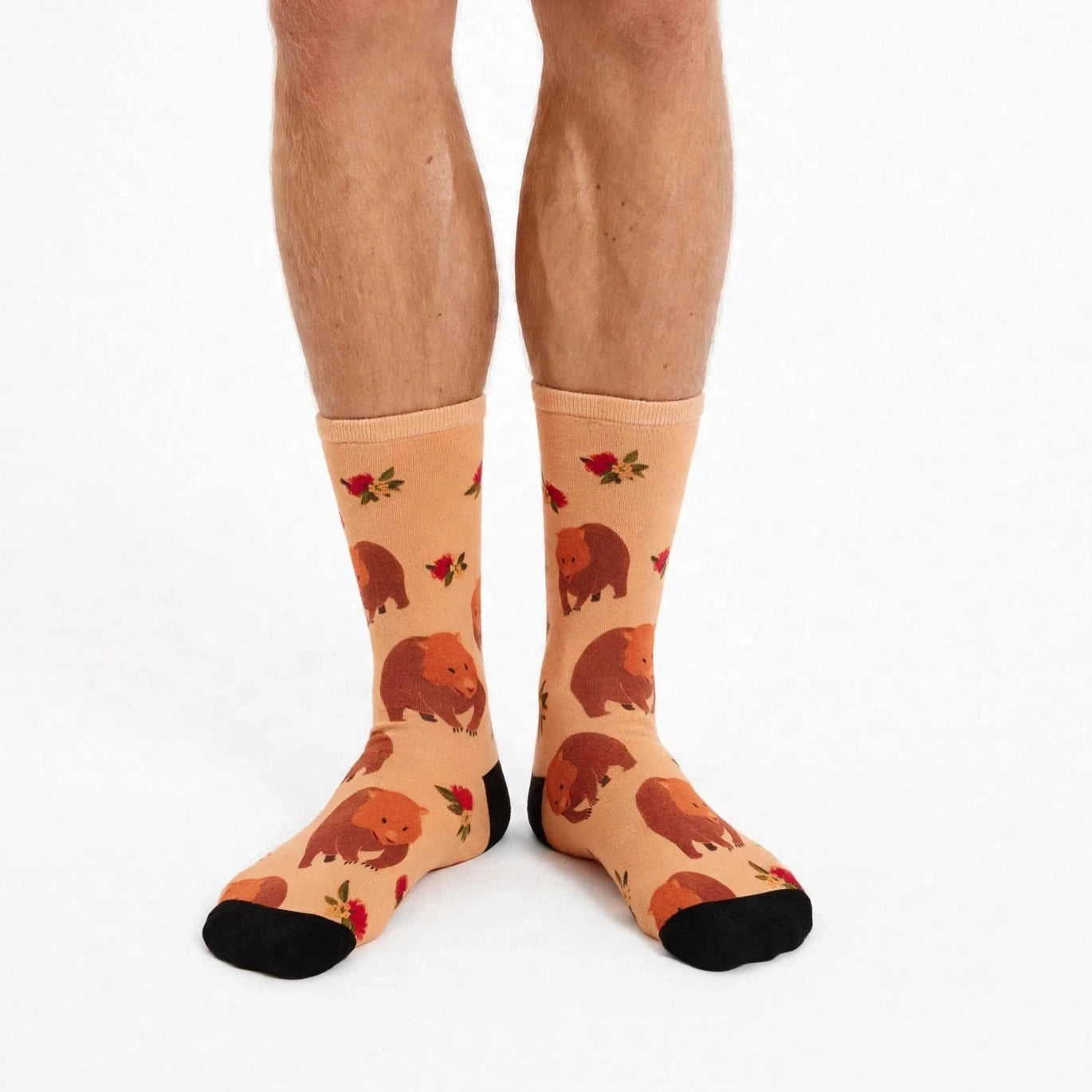 A pair of crew socks with a wombat and red rose pattern on a peach background, worn on a person's feet against a white studio background.
