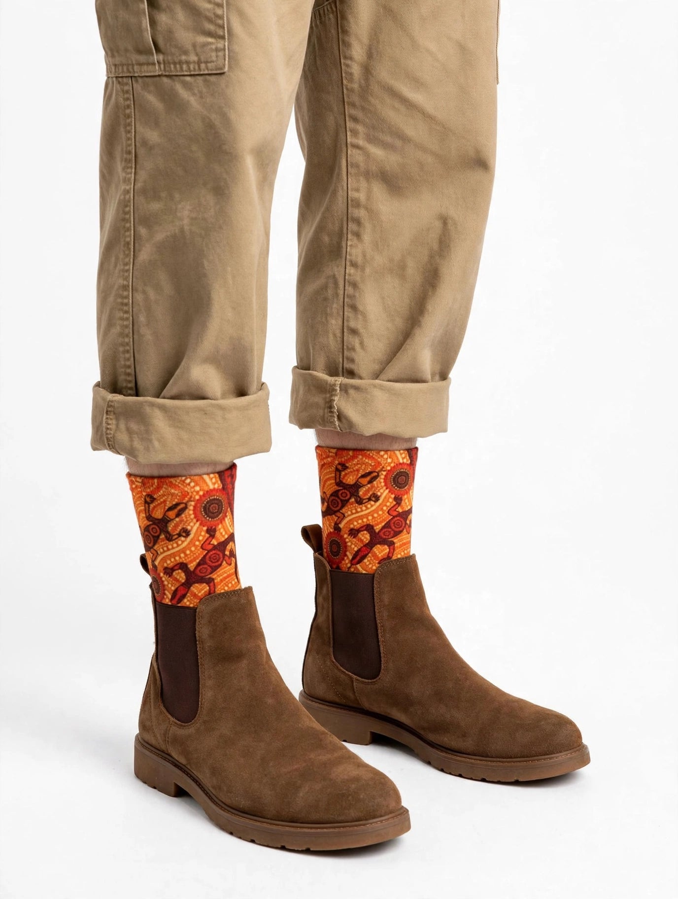 A person wearing brown suede Chelsea boots, cuffed tan pants, and bright orange Aboriginal patterned socks with a red and black design, shown against a plain white studio background.