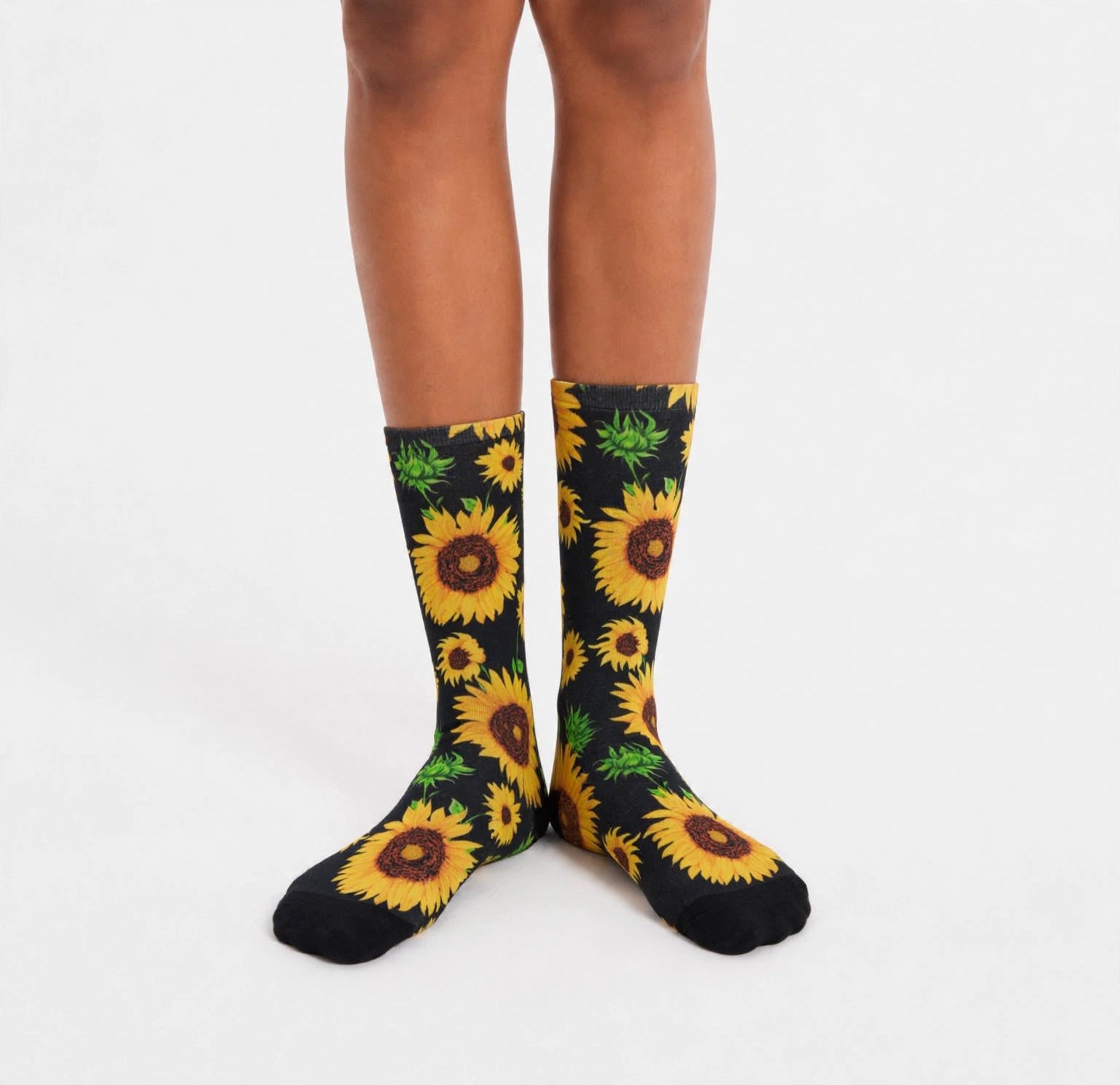 A person's legs wearing black crew socks with a vibrant yellow sunflower and green leaf pattern, photographed against a clean white studio background.