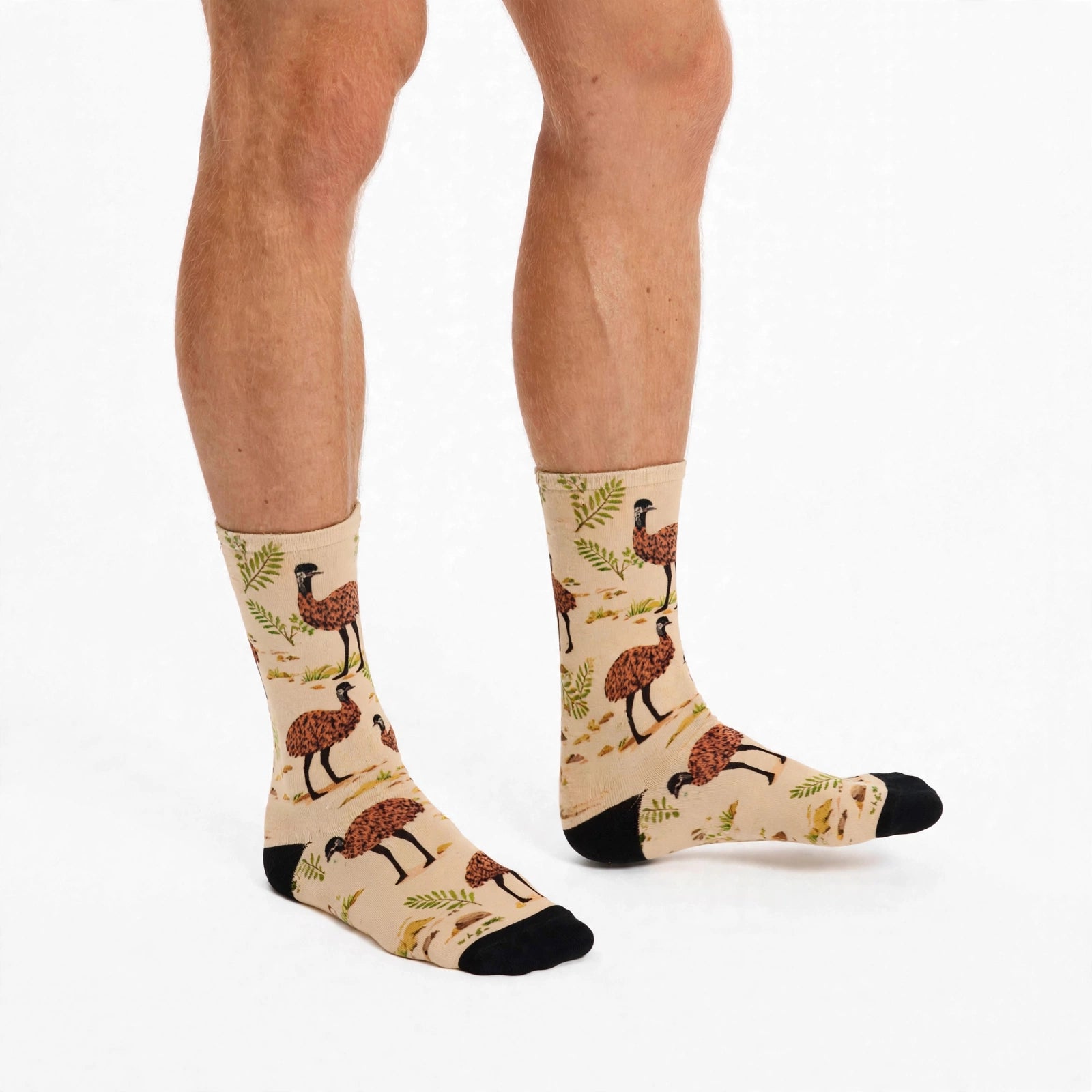 A pair of beige crew socks with a pattern of brown emus and green foliage, worn on a person's feet against a plain white background.