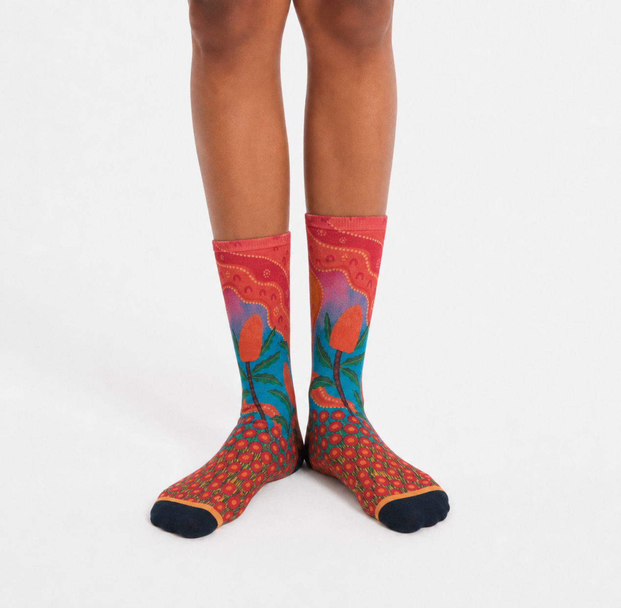 Colourful Aboriginal art socks featuring orange banksia flowers, native plant details and warm sunset-inspired colours, designed by Caitlin Trindall.