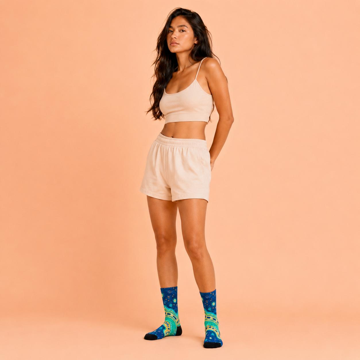 A woman modeling a matching beige crop top and shorts set, paired with blue socks featuring Aboriginal art, set against a solid peach studio background.