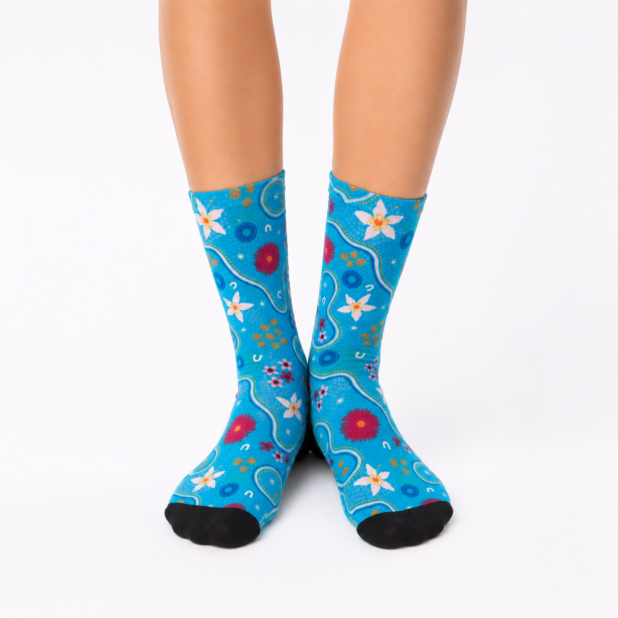 A pair of crew socks with a bright blue background and a colorful floral and Aboriginal  pattern, worn on a person's feet against a light gray studio background.