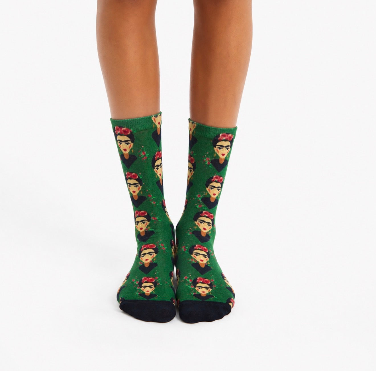 A pair of green crew socks with a repeating print of Frida Kahlo's face and black toes, worn on a model's feet against a plain white background.