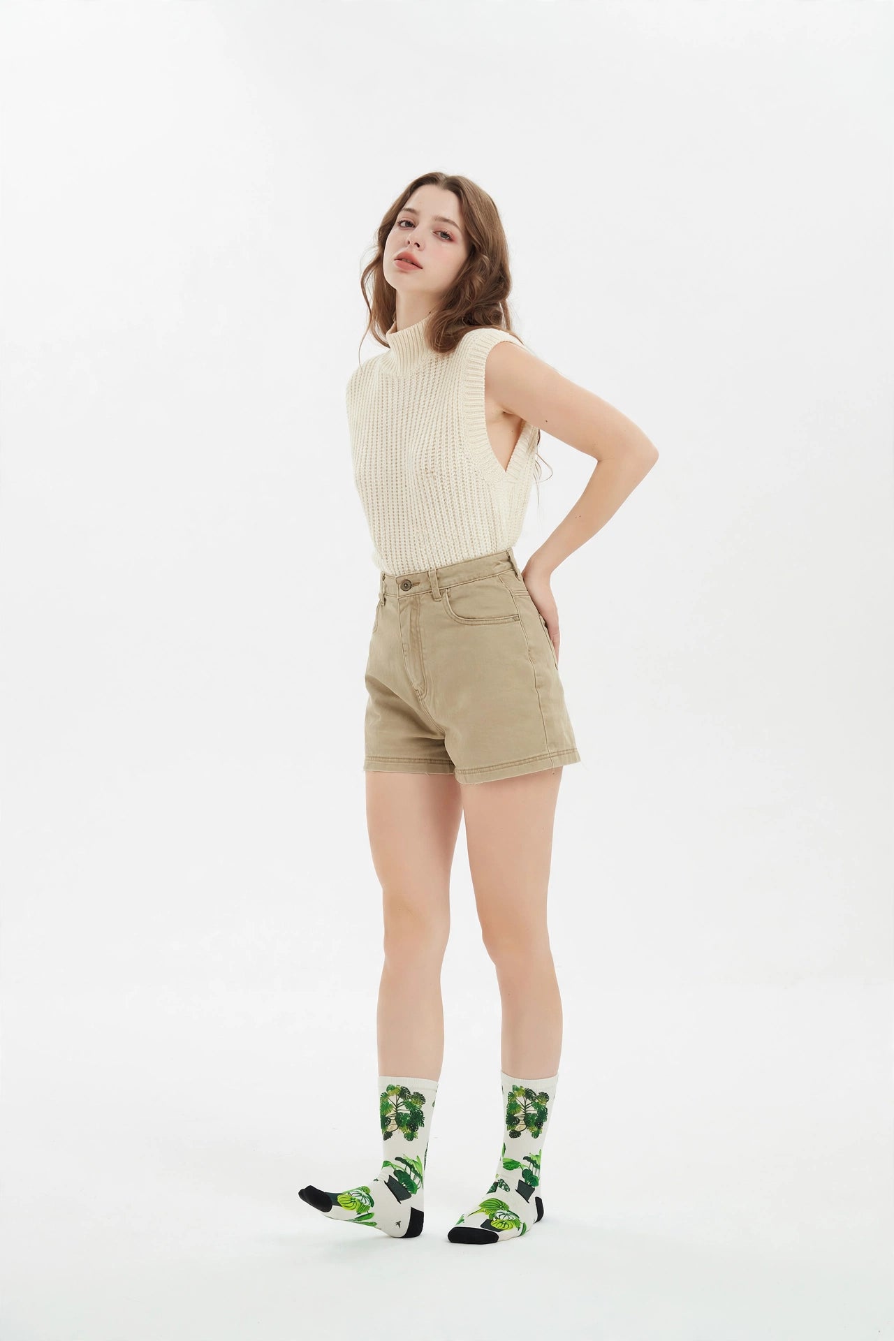 A woman modeling a cream ribbed sleeveless top, beige shorts, and white crew socks with a green and black pattern, standing against a plain white studio background.