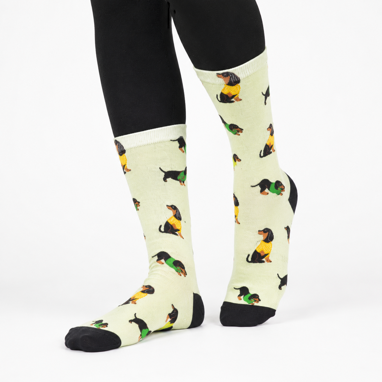 A pair of knee-high socks with a repeating pattern of dachshund dogs wearing yellow and green sweaters, modeled on a person's legs wearing black leggings against a plain white background.