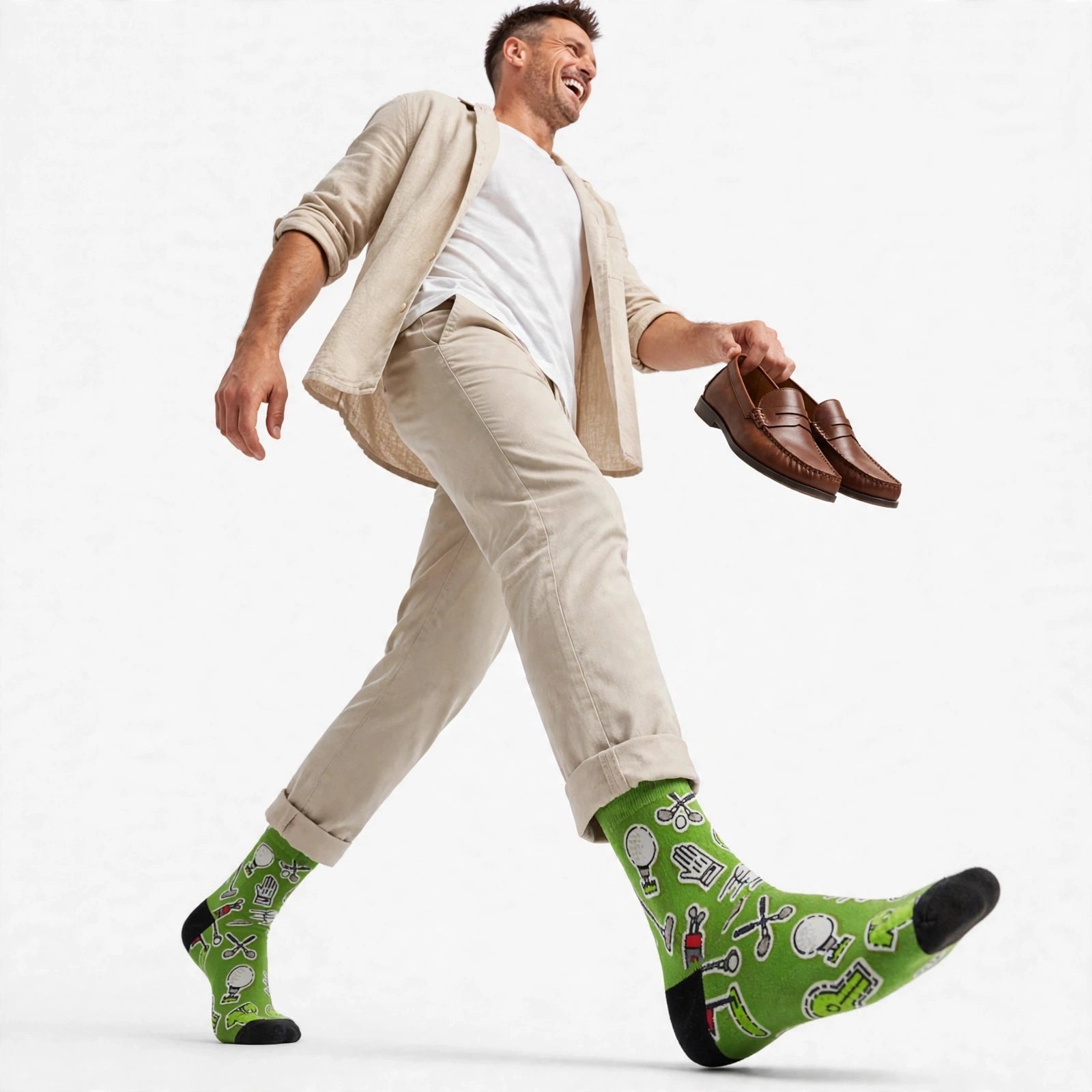 A man walking in a studio wearing bright green novelty golf socks with cartoon illustrations, a beige blazer, white t-shirt, and beige trousers, while holding a pair of brown leather shoes.