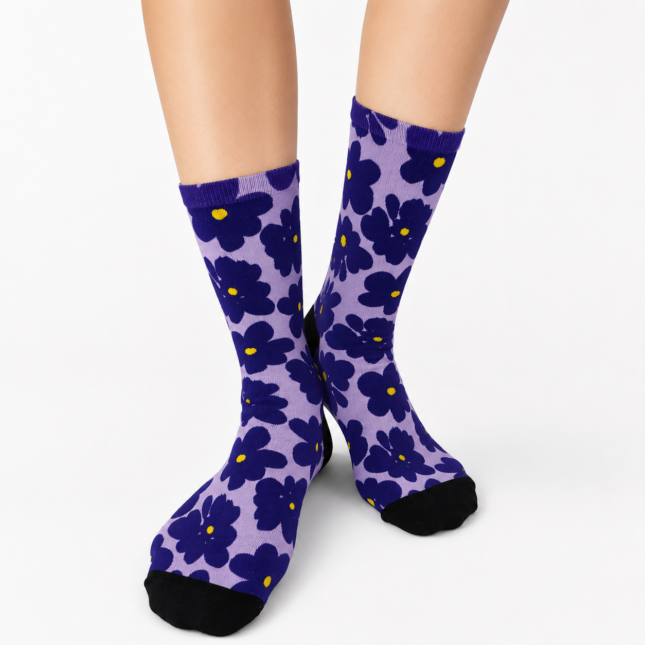 A pair of crew socks with a purple and dark blue floral pattern, featuring small yellow flower centers and black toes and heels, shown on a model's feet against a white background.
