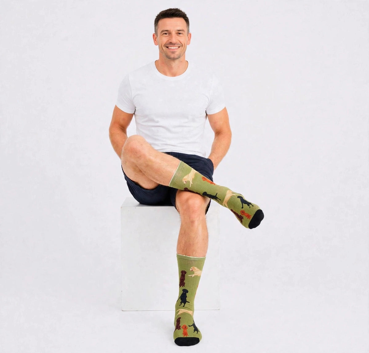 A man sitting on a white block wearing olive green Labrador socks with a camel and rider pattern, a white t-shirt, and navy shorts against a light gray background.