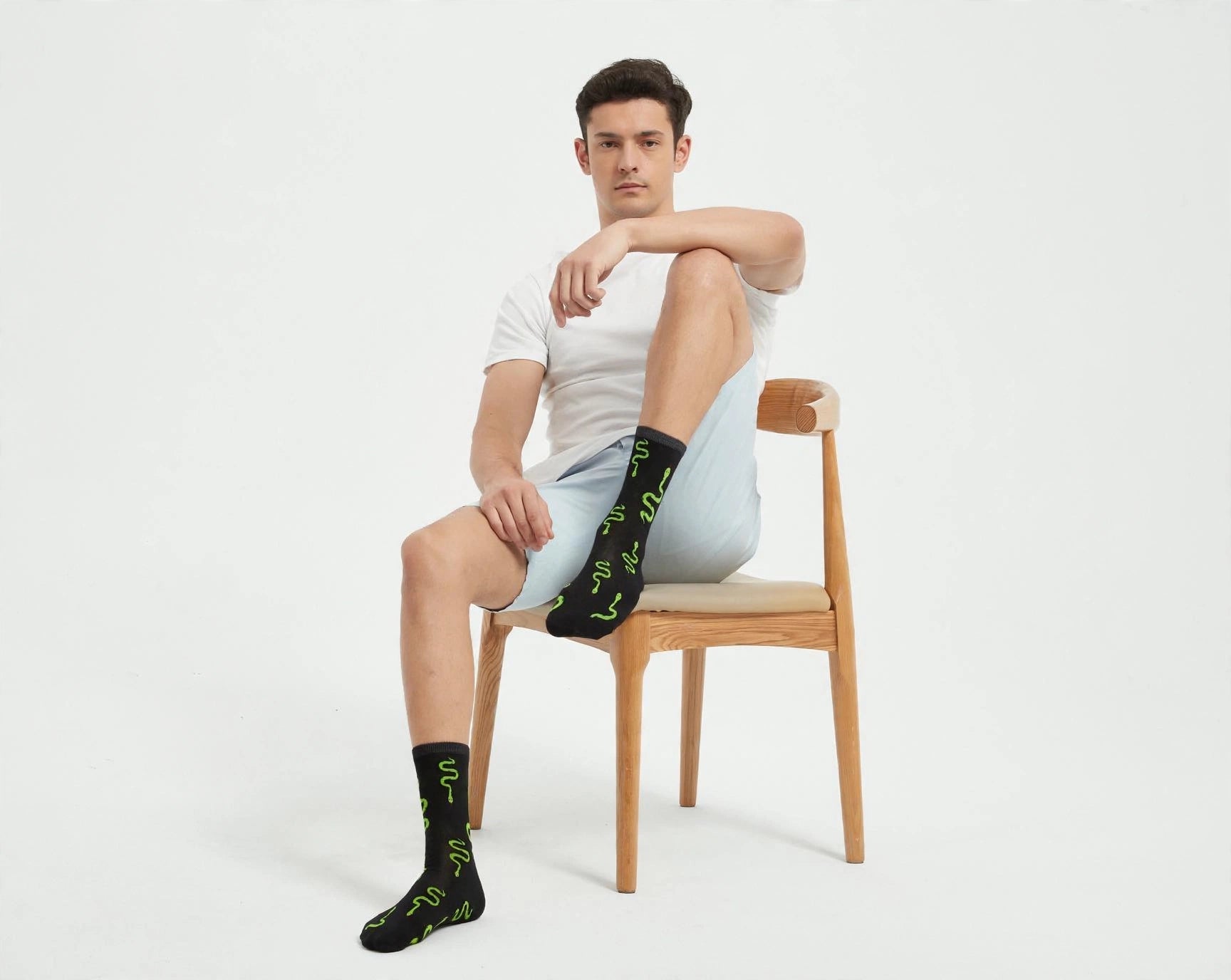 A man sits on a wooden chair wearing a white t-shirt, light blue shorts, and black crew socks with a green snake pattern, against a plain white studio background.