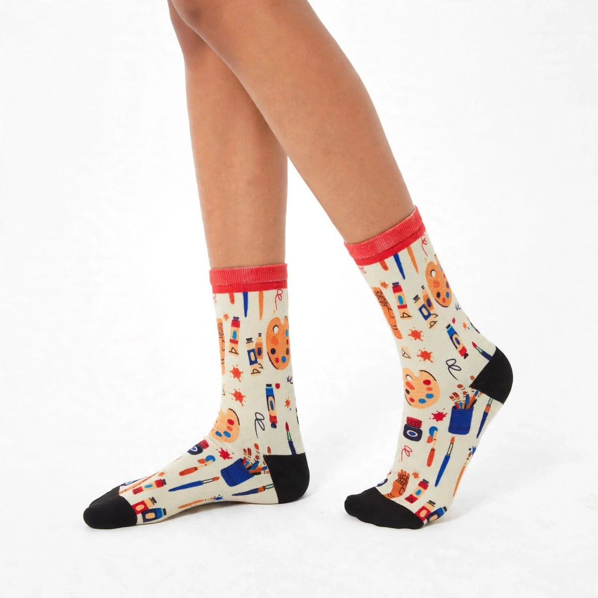 A pair of crew socks with a colorful artist-themed print on a cream background, featuring red cuffs and black toes/heels, worn on a model's legs against a white studio background.