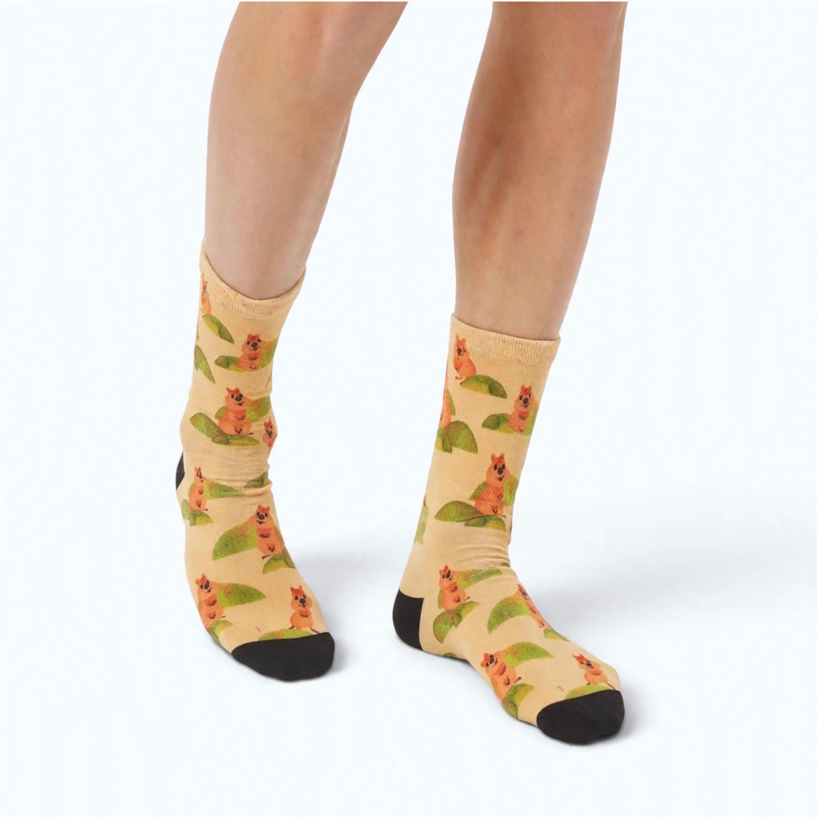 A person's legs wearing crew socks with an all-over print of quokka on a light beige background, featuring solid black toes and heels, photographed against a clean white studio background.