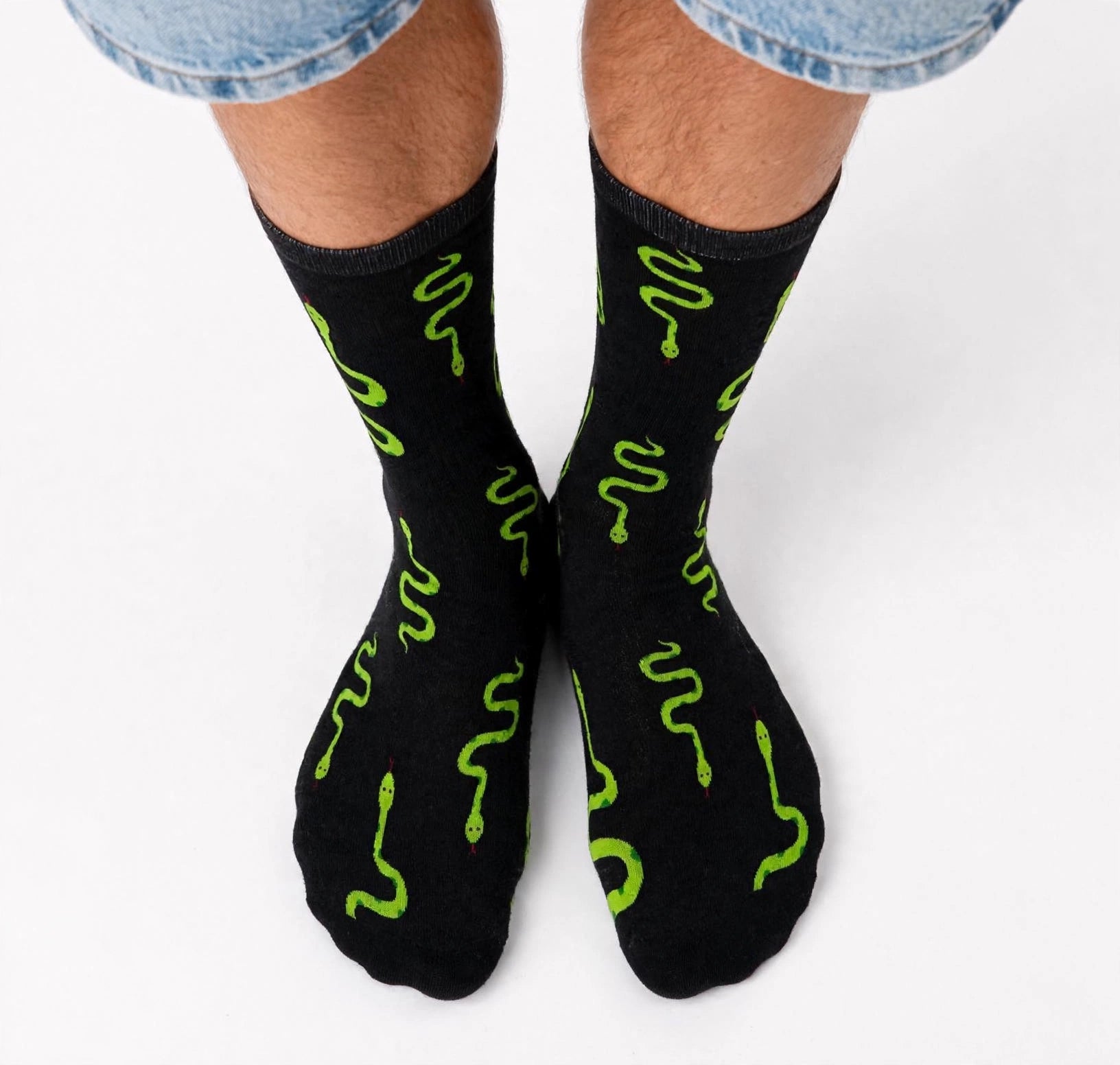 Black crew socks with a bright green snake print, worn by a model with blue denim shorts visible at the top, shot against a clean white background.