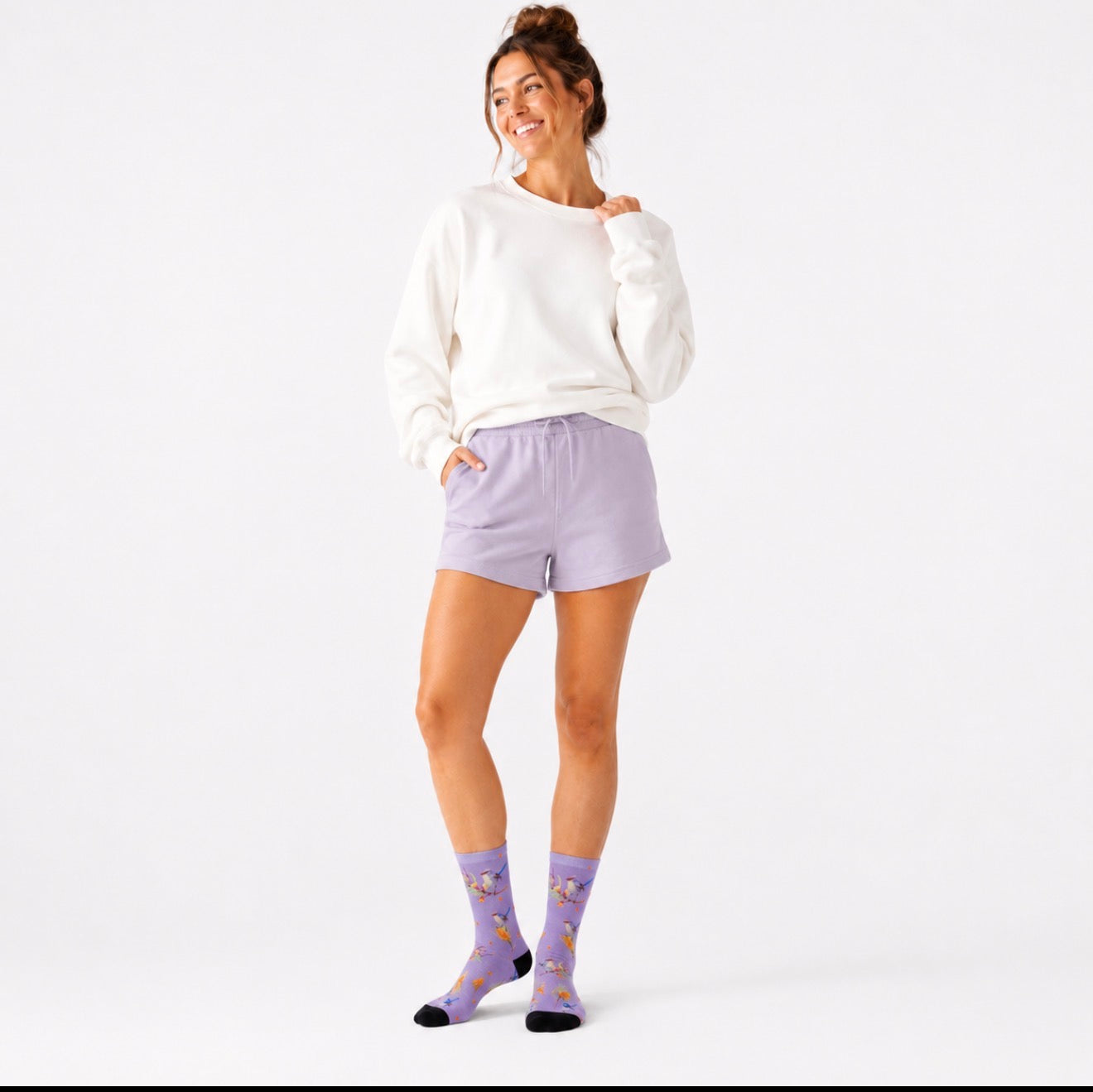 A woman modeling a white crewneck sweatshirt, light purple shorts, and matching purple socks with a cartoon pattern, standing against a plain white studio background. Wearing Australia wren bird socks. 