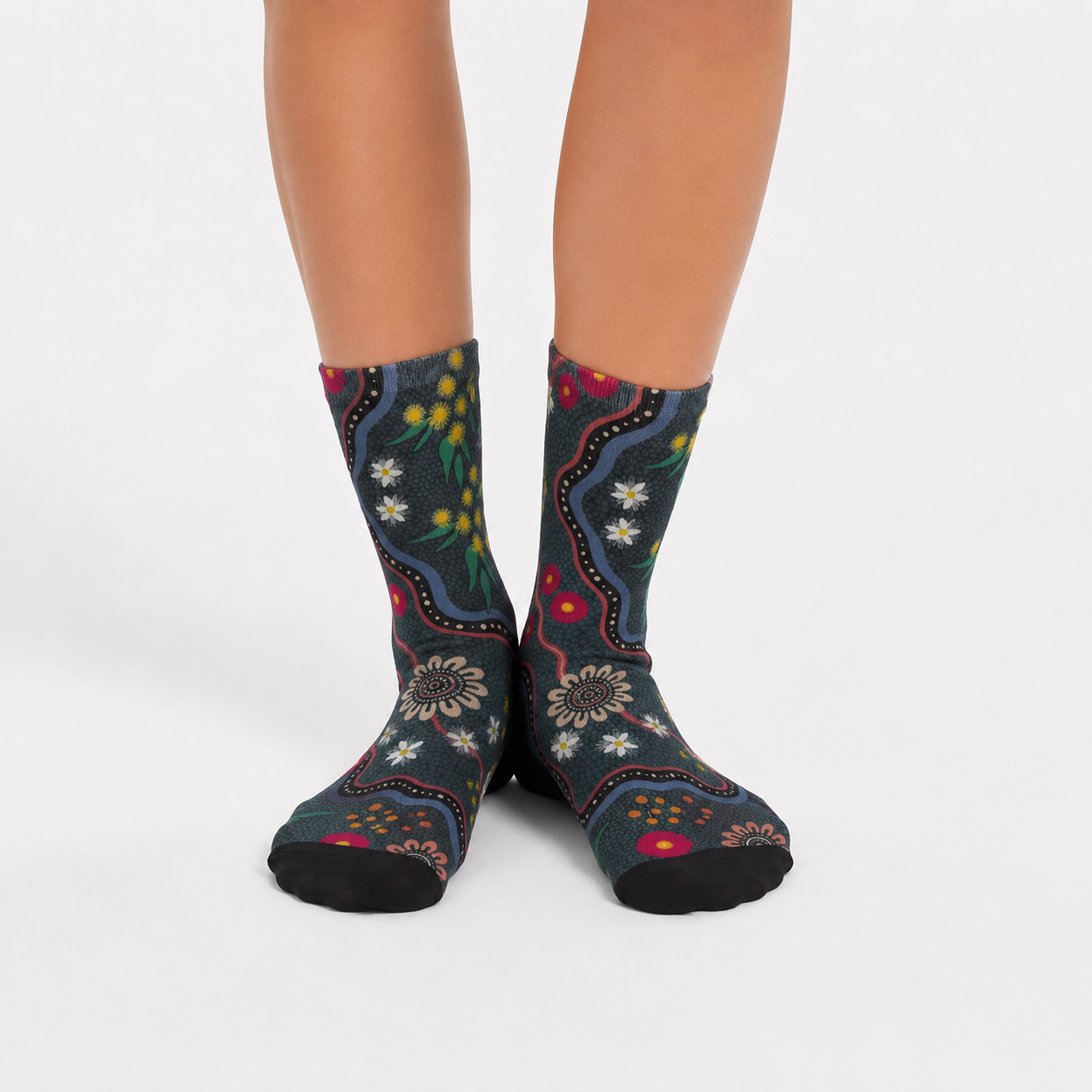 A pair of crew socks with a dark teal and black Aboriginal-inspired dot art pattern featuring red, yellow, green, and white details, worn on a person's feet against a light gray background.