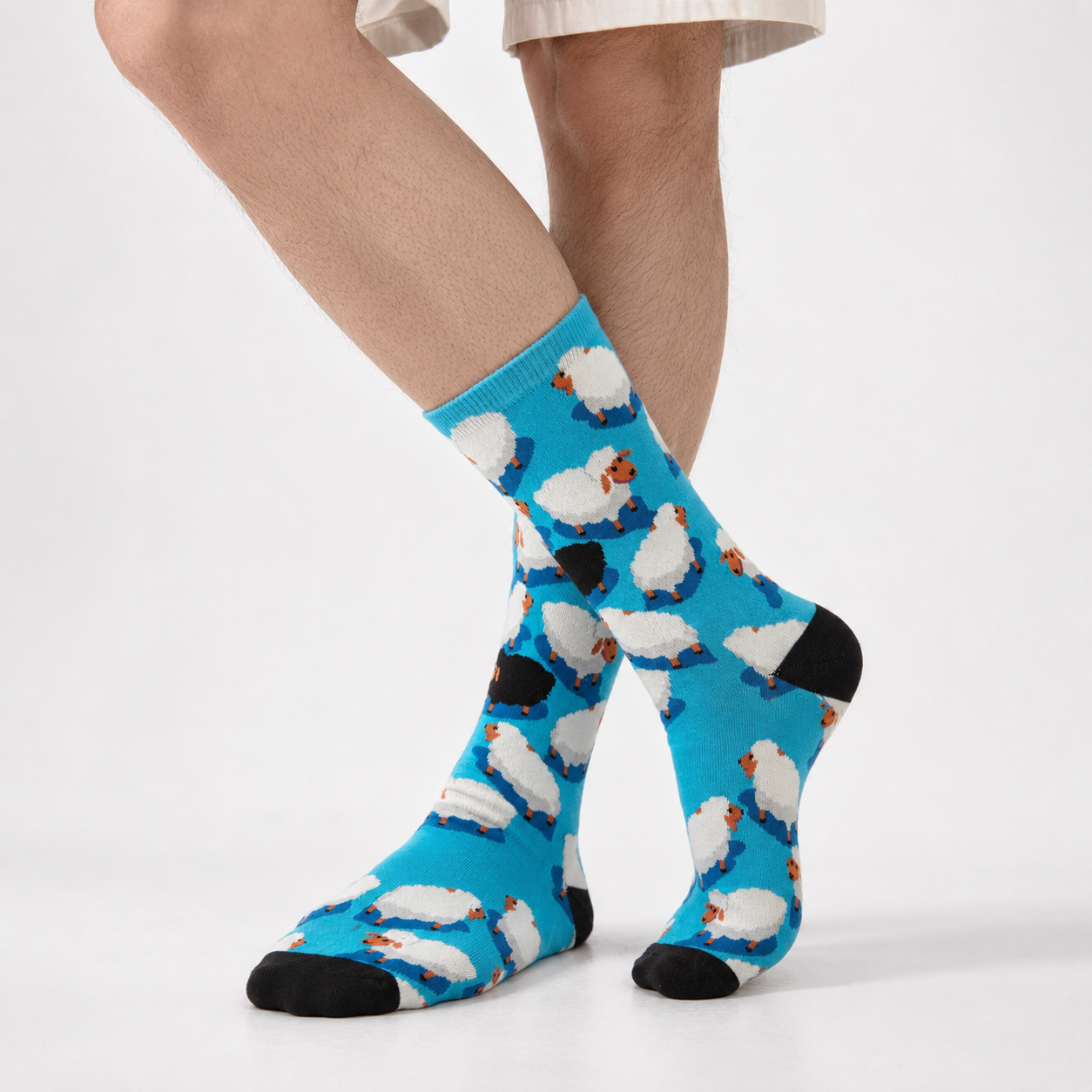 A person's legs wearing bright blue crew socks with a repeating white sheep pattern and black toe and heel accents, shown against a clean light gray studio background.