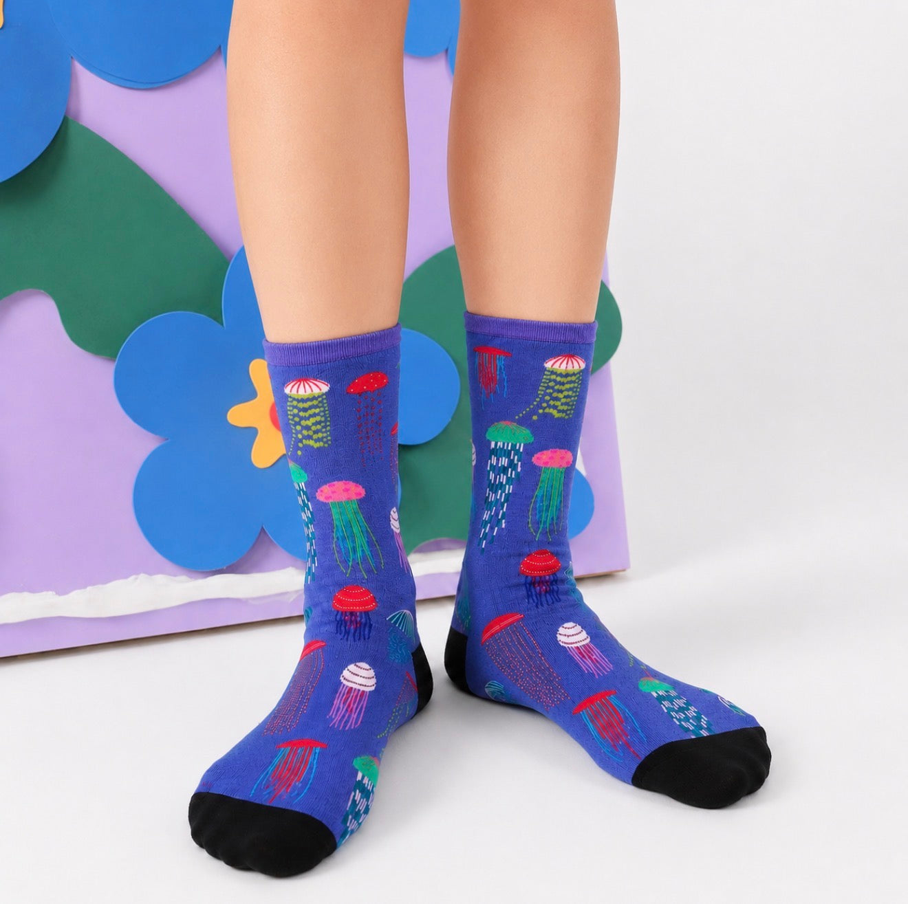 A person wearing blue crew socks with a colorful jellyfish pattern, standing in front of a decorative pastel backdrop.
